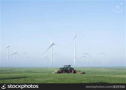 farmer mows grass near wind turbine farm in oastfriesland on misty summer morning in german ostfriesland