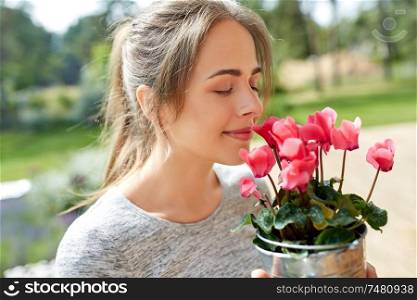 farming, gardening and people concept - happy young woman smelling pink cyclamen flowers in pot at summer garden. young woman with cyclamen flowers at summer garden