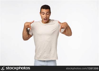 Fascinated and astonished, handsome speechless hispanic muscular man in white t-shirt, holding breath from fascination and admiration, stare surprised down, pointing bottom promo, white background.. Fascinated and astonished, handsome speechless hispanic muscular man in white t-shirt, holding breath from fascination and admiration, stare surprised down, pointing bottom promo, white background
