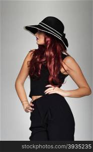 Fashion and beauty model in a chic black ensemble. Fashion and beauty model with lovely long curly auburn hair posing with her hands on her hips in a stylish black ensemble with matching wide brimmed black hat