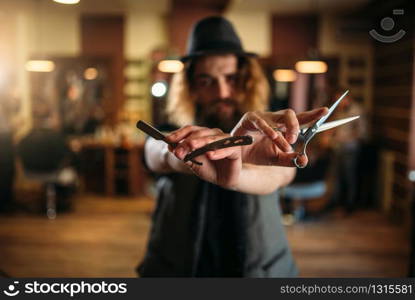 Fashion bearded barber in hat posing with scissors and straight razor in hands, selective focus, blur background