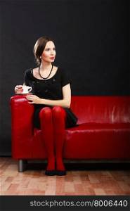Fashion beauty and elegance concept. Woman retro style in full length. Elegant lady holding coffee tea cup hot drink sitting on red sofa, indoor