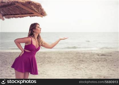Fashion blonde portrait happy at the beach posing on sand under tiki patio umbrella dressed in a fuchsia pink violet dress. Holding hand, Copy space on the right