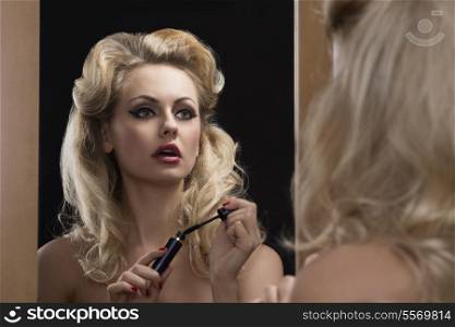 fashion blonde woman with elegant hair-style and make-up applying mascara in front of the mirror