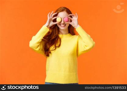 Fashion, food and consumer concept. Cheerful silly redhead woman fooling around, playing with dessert, make eyes from macarons and smiling joyfully, standing orange background.. Fashion, food and consumer concept. Cheerful silly redhead woman fooling around, playing with dessert, make eyes from macarons and smiling joyfully, standing orange background