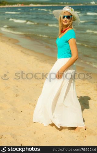 Fashion, happiness and lifestyle concept. Lovely blonde girl in full length in hat blue sunglasses and summer clothing walking on beach. Young woman relaxing on the sea coast.