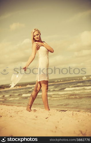 Fashion, happiness and lifestyle concept. Lovely blonde girl in full length wearing summer clothing lace white dress with hat in hand walking on beach. Young woman relaxing on the sea coast.