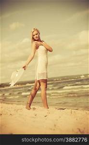 Fashion, happiness and lifestyle concept. Lovely blonde girl in full length wearing summer clothing lace white dress with hat in hand walking on beach. Young woman relaxing on the sea coast.