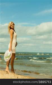 Fashion, happiness and lifestyle concept. Lovely blonde girl in full length wearing summer clothing lace white dress with hat in hand walking on beach. Young woman relaxing on the sea coast.