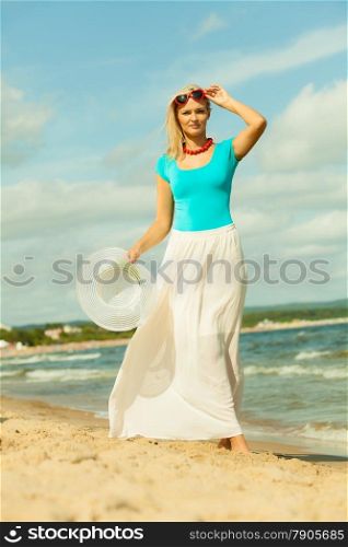 Fashion, happiness and lifestyle concept. Lovely blonde girl in full length with hat in hand red sunglasses and summer clothing walking on beach. Young woman relaxing on the sea shore.