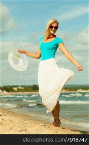 Fashion, happiness and lifestyle concept. Lovely blonde girl in full length with hat in hand red sunglasses and summer clothing walking on beach. Young woman relaxing on the sea shore.