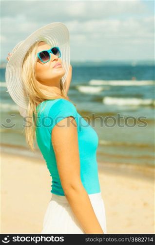 Fashion, happiness and lifestyle concept. Lovely blonde girl in hat and blue sunglasses walking on beach. Young woman relaxing on the sea coast.