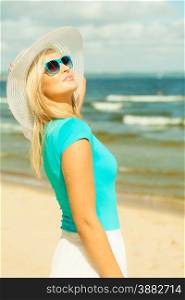 Fashion, happiness and lifestyle concept. Lovely blonde girl in hat and blue sunglasses walking on beach. Young woman relaxing on the sea coast.