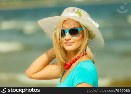 Fashion, happiness and lifestyle concept. Lovely blonde girl in hat and blue sunglasses red beads walking on beach. Young woman relaxing on the sea coast.