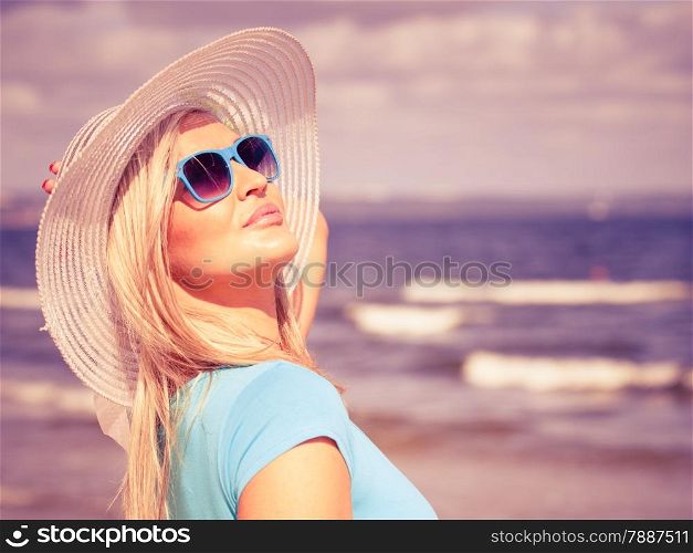 Fashion, happiness and lifestyle concept. Lovely blonde girl in hat and color sunglasses walking on beach. Young woman relaxing on the sea coast.