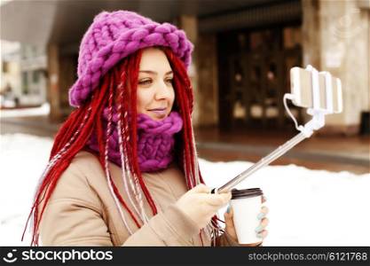 Fashion lifestyle portrait of young lovely woman wearing a trendy cap, a bright youth clothing, and making selfie with a stick against city background. Having fun, joy and happiness.