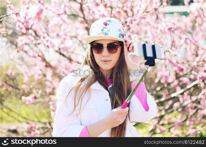 Fashion lifestyle portrait of young lovely woman wearing a trendy cap, a bright youth clothing, and making selfie with a stick against pink spring flowers. Having fun, joy and happiness.