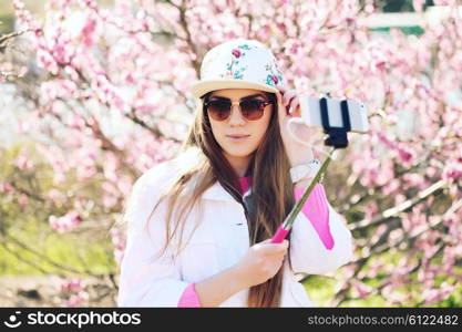 Fashion lifestyle portrait of young lovely woman wearing a trendy cap, a bright youth clothing, and making selfie with a stick against pink spring flowers. Having fun, joy and happiness.