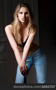 fashion low key shot of beautiful hot blond woman in black bra lingerie and jeans on dark indoor backgound