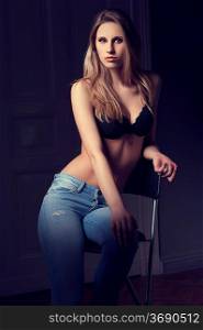 fashion low key shot of hot blond woman in black bra lingerie and jeans on dark indoor backgound