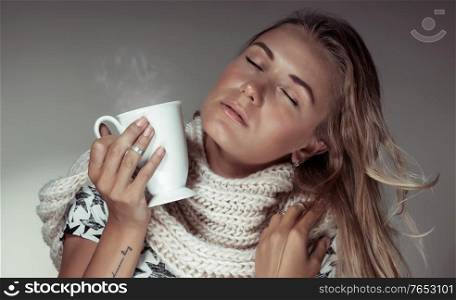 Fashion portrait of a nice blond woman with hot cup of coffe in hands wearing warm knitted scarf and closing eyes of pleasure, autumn season concept