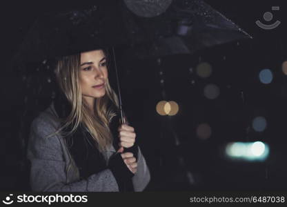 Fashion portrait of beautiful gorgeous woman standing with umbrella underthe rain over blury night city lights background, melancholy concept. Lonely woman at night