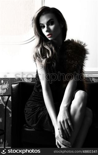 fashion portrait of beautiful woman with blue elegant shiny dress and fur on shoulders. Sitting on sofa in elegant ambient and looking in camera , black and white image with high contrast