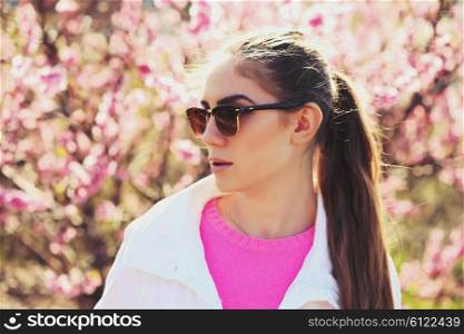 Fashion portrait of gorgeous young sexy blond girl posing in a lush garden in the spring in a stylish white jacket, a pink blouse and striped skirt and sunglasses. Bright young outfit.