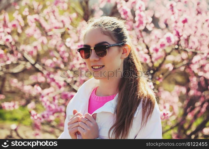 Fashion portrait of gorgeous young sexy blond girl posing in a lush garden in the spring in a stylish white jacket, a pink blouse and striped skirt and sunglasses. Bright young outfit.