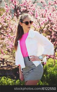Fashion portrait of gorgeous young sexy blond girl posing in a lush garden in the spring in a stylish white jacket, a pink blouse and striped skirt and sunglasses. Bright young outfit.