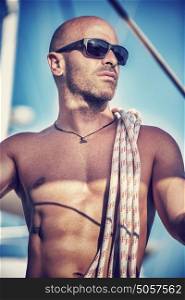 Fashion portrait of handsome shirtless man standing on the deck of sailboat with rope on shoulders in bright sun lights, sexy sailor, summer vacation and holidays