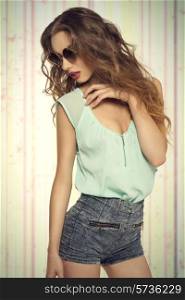 fashion portrait of lovely trendy brunette girl with red lipstick and long curly hair posing with sexy slim body wearing denim shorts, blue shirt and hipster sunglasses