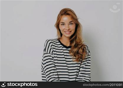 Fashion portrait of relaxed positive young woman in casual striped longsleeve with happy smile looking at camera, feeling charming while posing isolated over grey studio wall background. Relaxed positive young woman in casual striped longsleeve with happy smile looking at camera