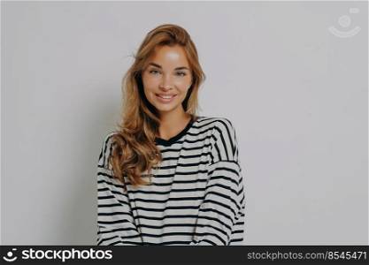 Fashion portrait of relaxed positive young woman in casual striped longsleeve with happy smile looking at camera, feeling charming while posing isolated over grey studio wall background. Relaxed positive young woman in casual striped longsleeve with happy smile looking at camera