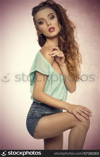 fashion portrait of sensual young woman in sexy pose with her perfect body wearing denim shorts, blue shirt and vintage sunglasses on the head. Stylish make-up, charming expression