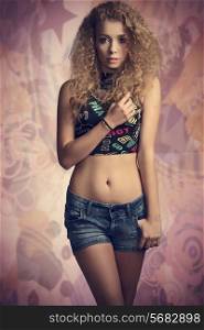 fashion portrait of very pretty teenager girl with curly hair and sexy casual clothes, posing with trendy top and denim shorts, looking in camera with innocent expression
