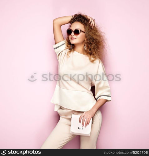 Fashion portrait of woman in summer outfit. Girl posing on pink background. Pink handbag and sunglasses. Stylish curly hairstyle. Glamour lady