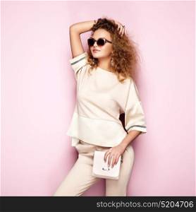 Fashion portrait of woman in summer outfit. Girl posing on pink background. Pink handbag and sunglasses. Stylish curly hairstyle. Glamour lady
