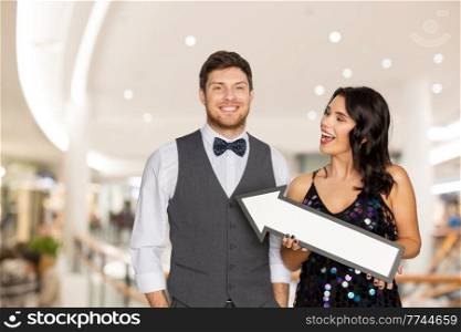 fashion, sale and consumerism concept - happy couple with big arrow at party over shopping mall background. happy couple with big arrow over shopping mall