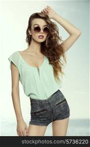 fashion shoot of sensual young model with long natural curly hair, she posing with vintage sunglasses, blue shirt and sexy denim shorts.
