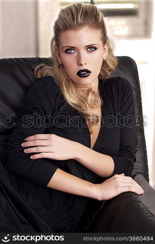 fashion shot of a sophisticated blonde sitting on a black sofa wearing a black dress and black lipstick