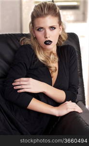 fashion shot of a sophisticated blonde sitting on a black sofa wearing a black dress and black lipstick