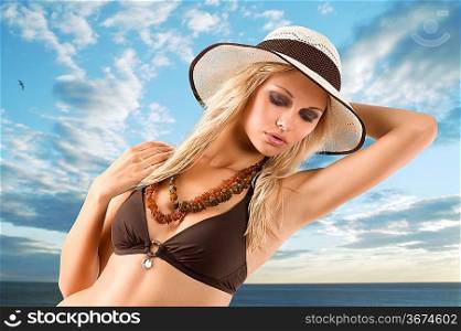 fashion shot of blond young girl in bikini swimsuit posing with hat against a cloudy sky