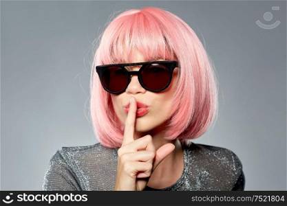 fashion, silence and secret concept - young woman in pink wig and black sunglasses making hush gesture over grey background. happy woman in pink wig and black sunglasses