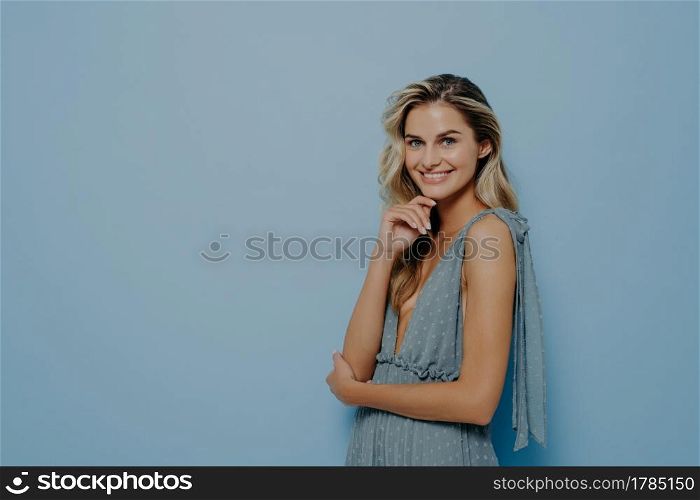 Fashion studio photo of beautiful woman with blond wavy hair and natural makeup, wears delicate party dress, posing sideways with folded hands and gently touching her face with fingers, softly smiling. Fashion studio photo of beautiful woman with blond wavy hair wears party dress softly smiling