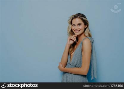 Fashion studio photo of beautiful woman with blond wavy hair and natural makeup, wears delicate party dress, posing sideways with folded hands and gently touching her face with fingers, softly smiling. Fashion studio photo of beautiful woman with blond wavy hair wears party dress softly smiling