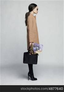 Fashion studio photo of young stylish woman. Beige coat, black leather boots and bag, bouquet of lavender. Catalogue clothes. Lookbook