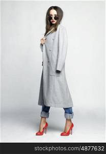 Fashion studio photo of young stylish woman. Grey oversize coat, white shirt, blue jeans, red shoes and handbag. Catalogue clothes. Lookbook