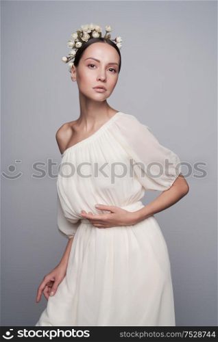 Fashion studio portrait of young beautiful woman in white greek dress with flowers on her head. Fashion and style