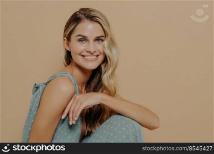 Fashion studio shot with smiling young woman on chair, charming pretty female in soft blue evening dress isolated over beige background with copy space, sitting sideways and looking at the camera. Fashion studio shot with smiling woman in evening dress sitting on chair